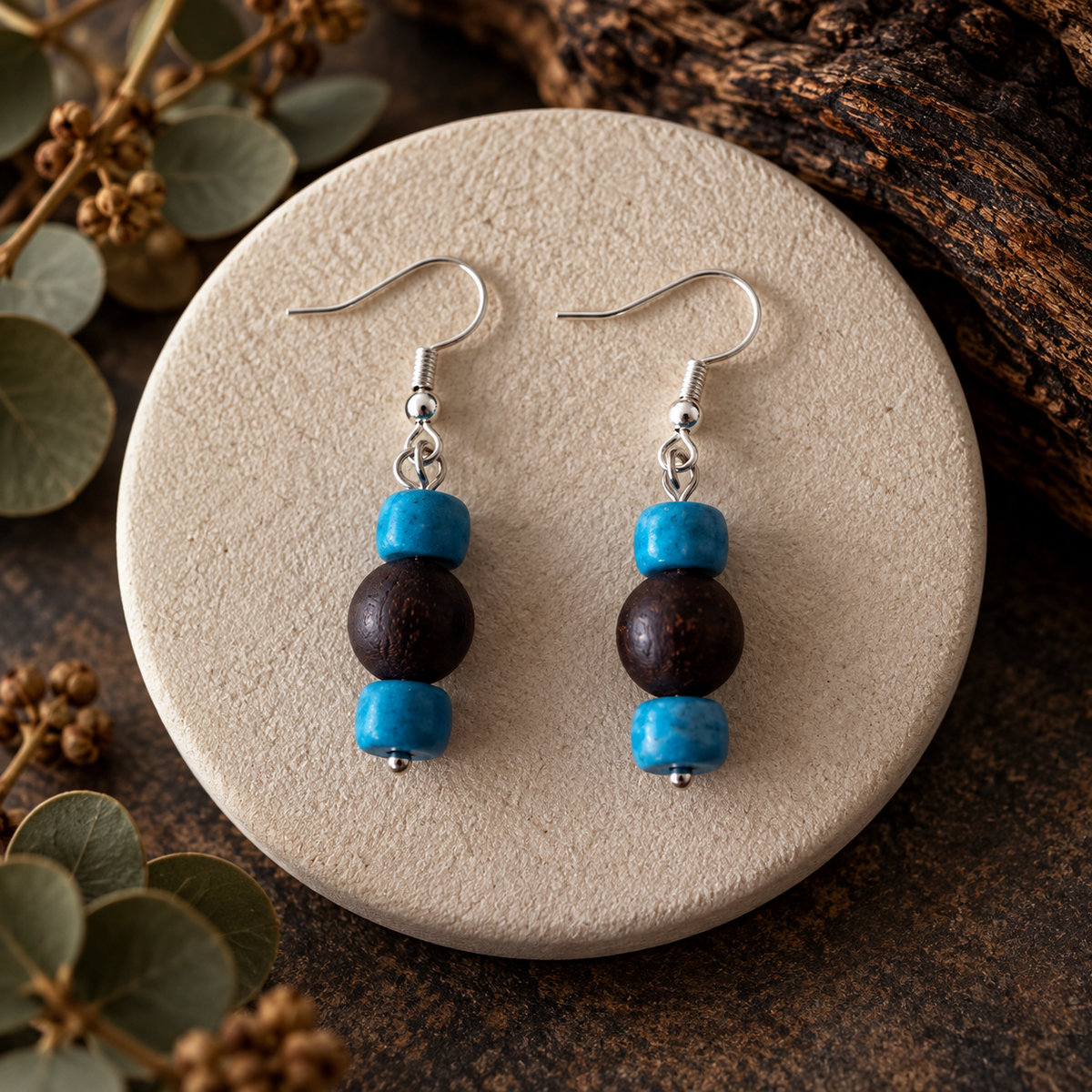 A pair of handmade dangle earrings with turquoise blue and dark brown wooden beads on silver fishhook wires, displayed on a round beige stone coaster with rustic wood and eucalyptus leaves in the background.