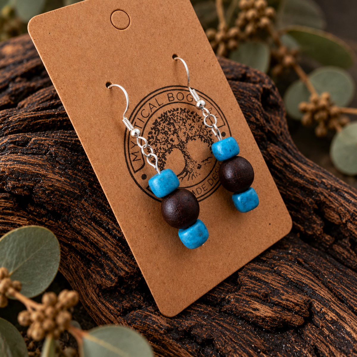 A pair of handmade dangle earrings displayed on a brown kraft paper card. The earrings feature silver-tone fishhook wires, a bright turquoise blue cylindrical bead, a dark brown wooden bead, and a smaller turquoise blue cylindrical bead.