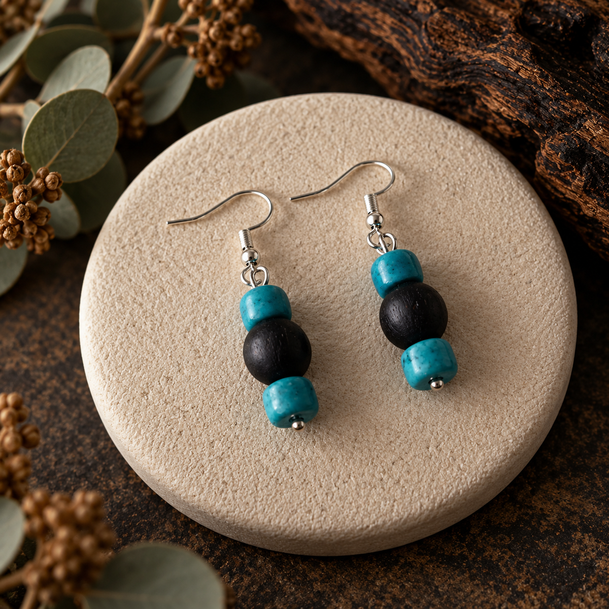 A pair of beaded dangle earrings displayed on a round beige stone coaster, with dried botanicals and dark wood in the background. The earrings feature silver fishhook wires with three beads: two glossy black round beads and one turquoise blue oval bead at the bottom.