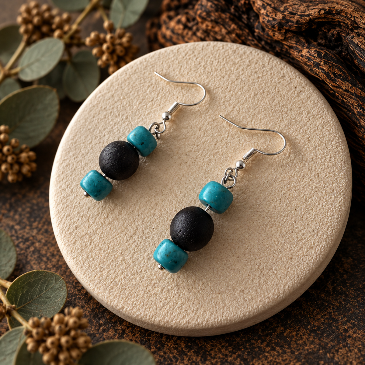 A pair of dangle earrings displayed on a round beige stone tray, surrounded by eucalyptus leaves and dried flower buds. The earrings feature silver fishhook wires with a three-bead drop design: a large matte black round bead, a smaller textured turquoise blue cube bead, and a tiny round turquoise bead at the end.