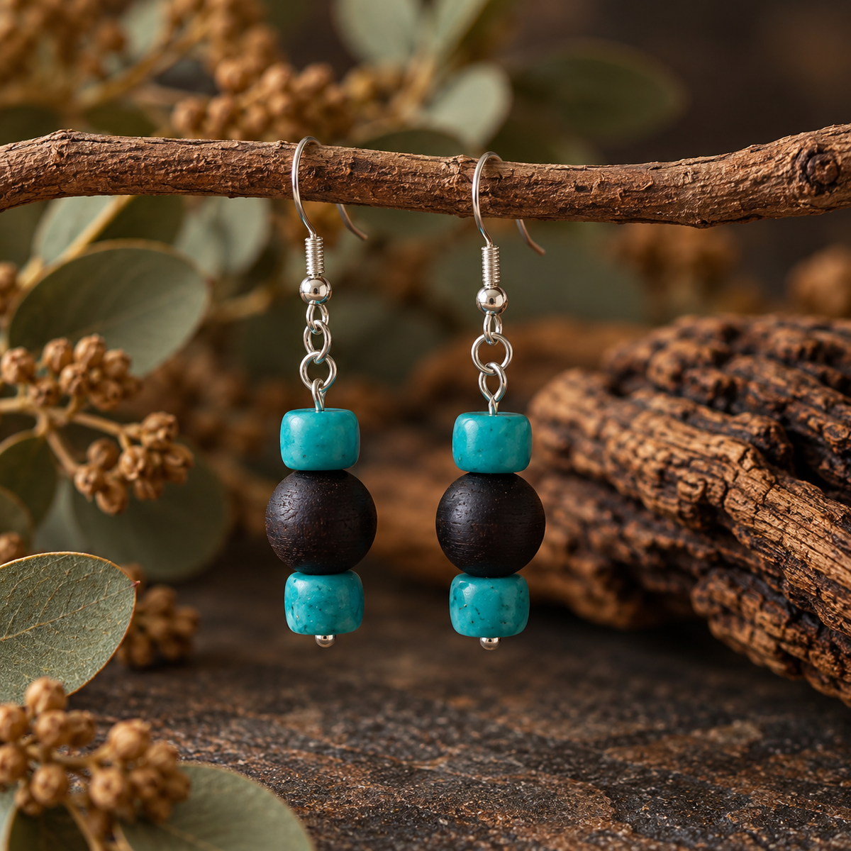 A pair of handmade dangle earrings hanging from a rustic branch. Each earring has a silver fishhook, a small silver bead, a turquoise blue disc bead, a large dark brown wooden bead, and a smaller turquoise blue disc bead at the bottom.