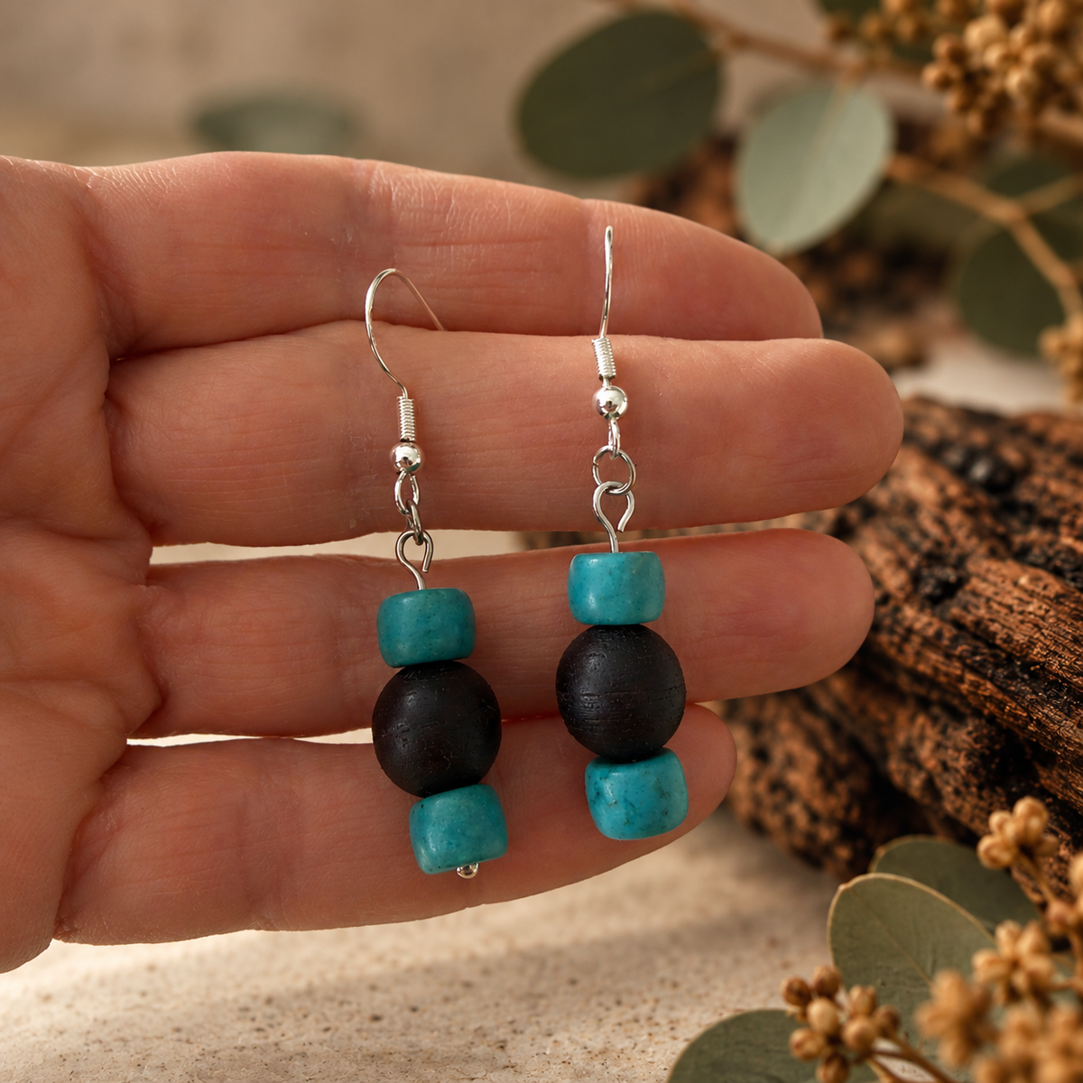 A pair of handmade dangle earrings held in a hand. The earrings feature silver fishhook wires with small silver beads, followed by a turquoise cube bead, a black round bead, and another turquoise cube bead at the bottom.