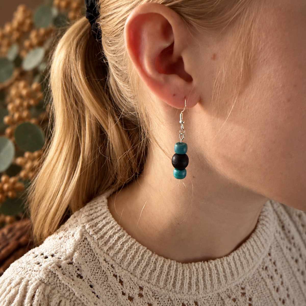 A close-up lifestyle shot of a woman wearing a silver dangle earring with two beads, one turquoise blue and one black, against a warm, softly blurred background.