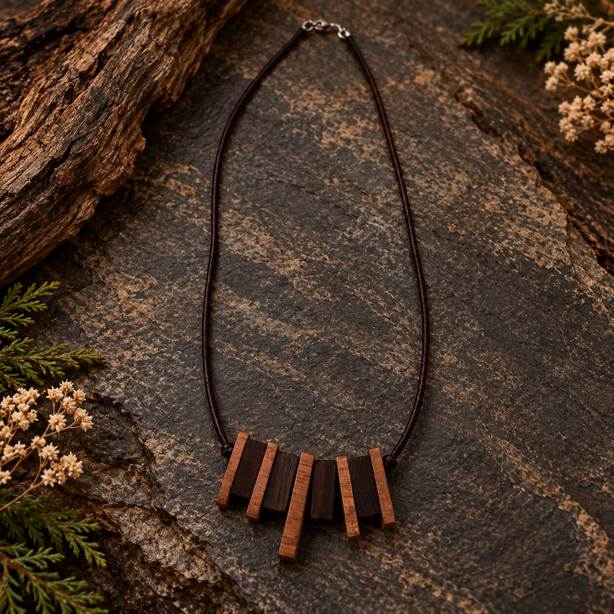 A dark brown cord necklace with a geometric pendant made of alternating dark and light brown wooden bars, displayed on a textured dark stone surface with natural wood and greenery props.