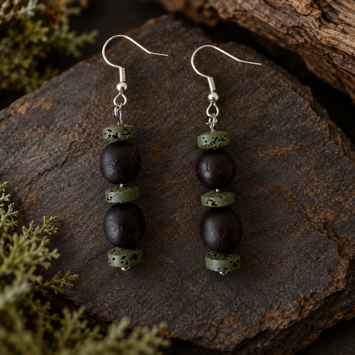 A pair of handmade dangle earrings with dark brown matte beads, light green speckled beads, and silver fishhook wires, displayed on a dark textured stone with moss and wood in the background.