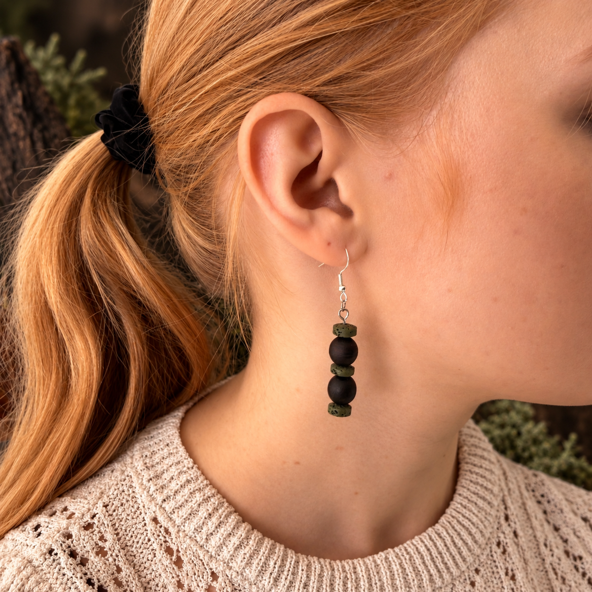 A close-up of a woman's ear wearing a dangling earring with four dark green beads and a silver fishhook wire, against her neck and hair.