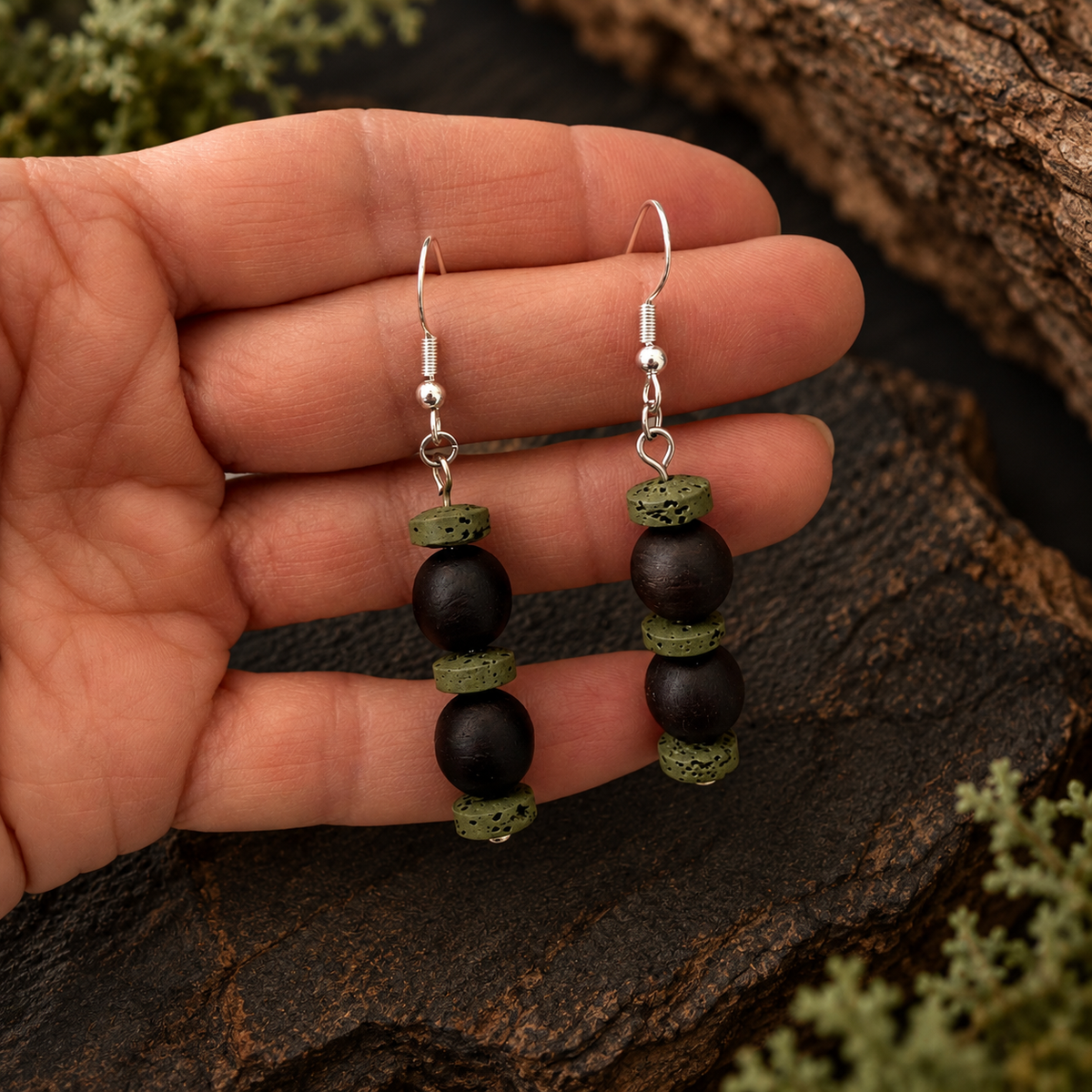 A pair of handmade dangle earrings held in a hand, featuring matte black beads, textured olive green beads, and silver fishhook wires, set against a rustic dark wood and moss background.