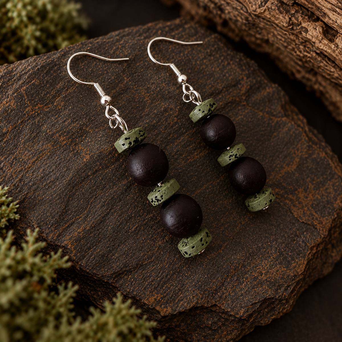 A pair of handmade dangle earrings with dark brown matte beads and textured sage green stone discs, hanging from silver fishhook wires on a rustic dark wood background.