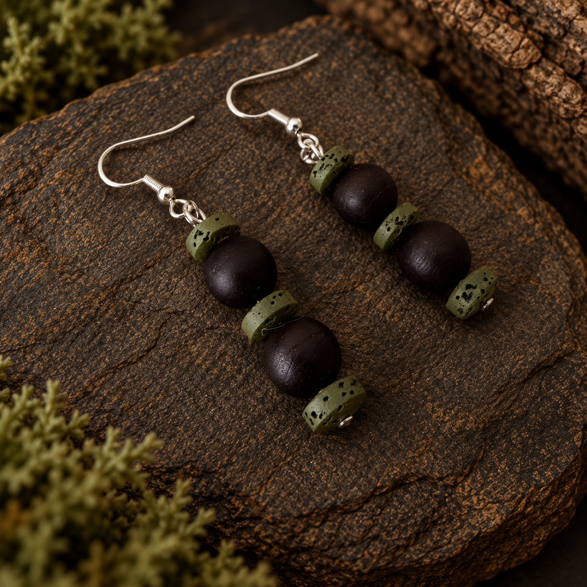 A pair of handmade dangle earrings with dark brown wooden beads and olive green stone spacers, displayed on a textured dark brown rock with moss.