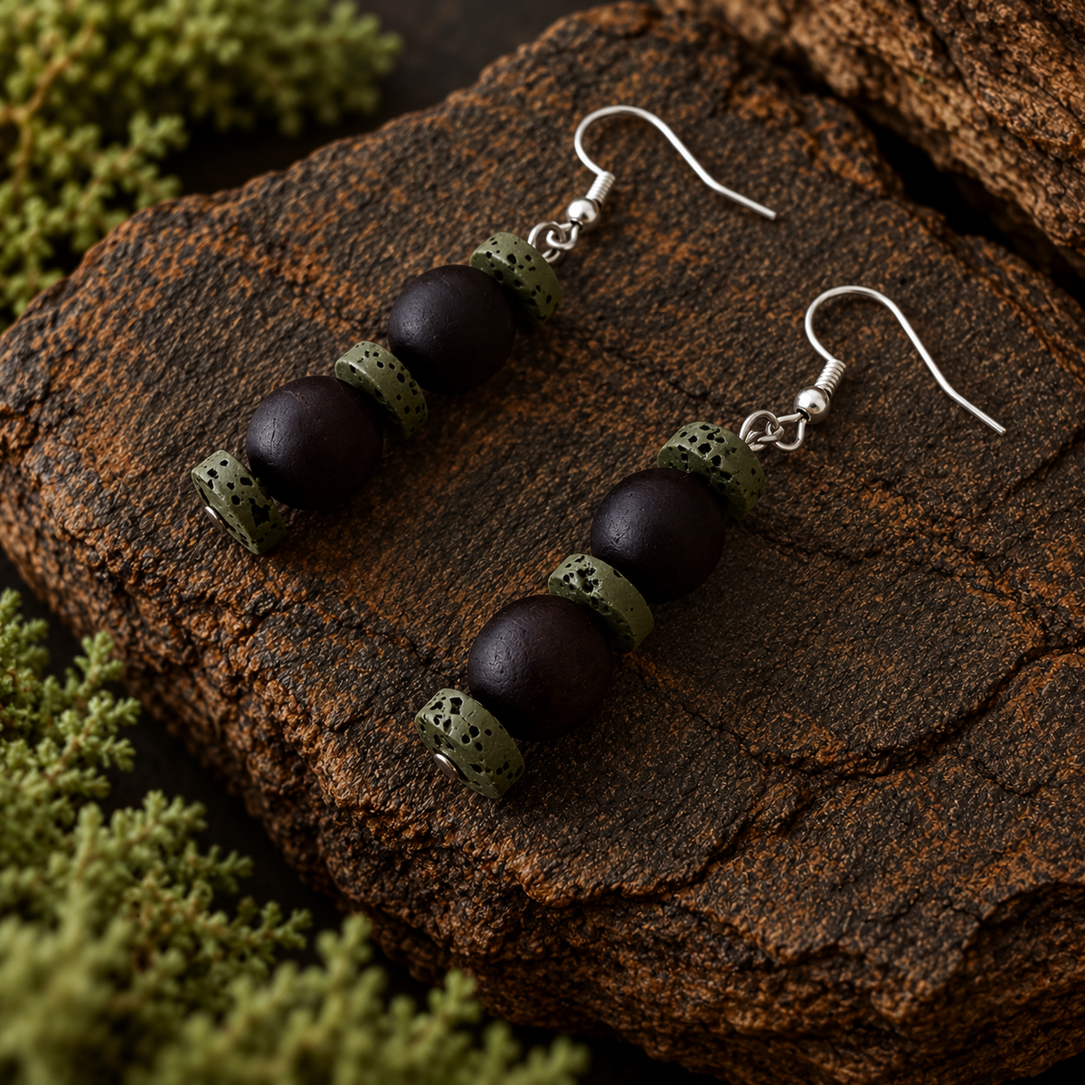 A pair of handmade dangle earrings with dark brown wooden beads and olive green perforated spacer beads, resting on a textured brown rock with green moss.