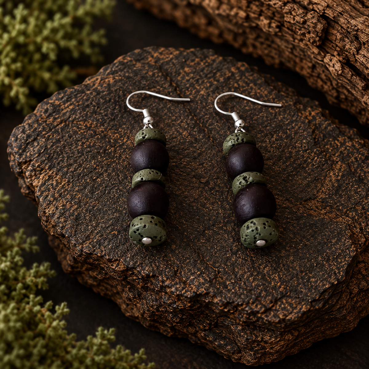 A pair of handmade dangle earrings with dark brown and olive green beads, displayed on a textured brown stone with natural moss and bark in the background.