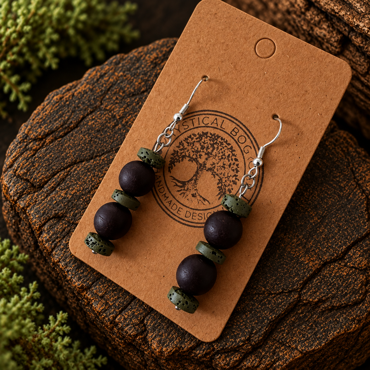 A pair of handmade dangle earrings displayed on a brown kraft card, which is placed on a textured wood log with green moss in the background. The earrings feature dark brown wooden beads and olive green stone spacers with silver-tone hooks.
