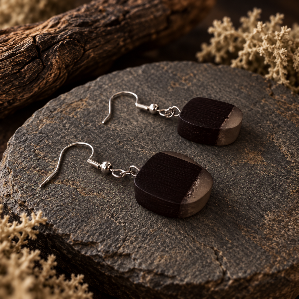 A pair of drop earrings with dark wood square pendants and silver fishhook wires, displayed on a rustic dark stone slab with dried botanicals and wood in the background.