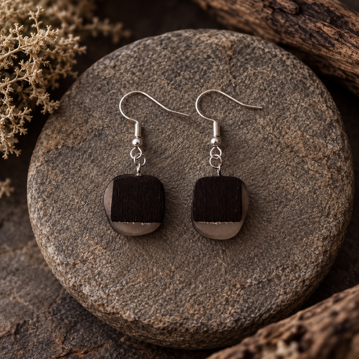 A pair of handmade dangle earrings with square dark brown wooden pendants and silver fishhook wires, displayed on a round gray stone with dried botanicals in the background.