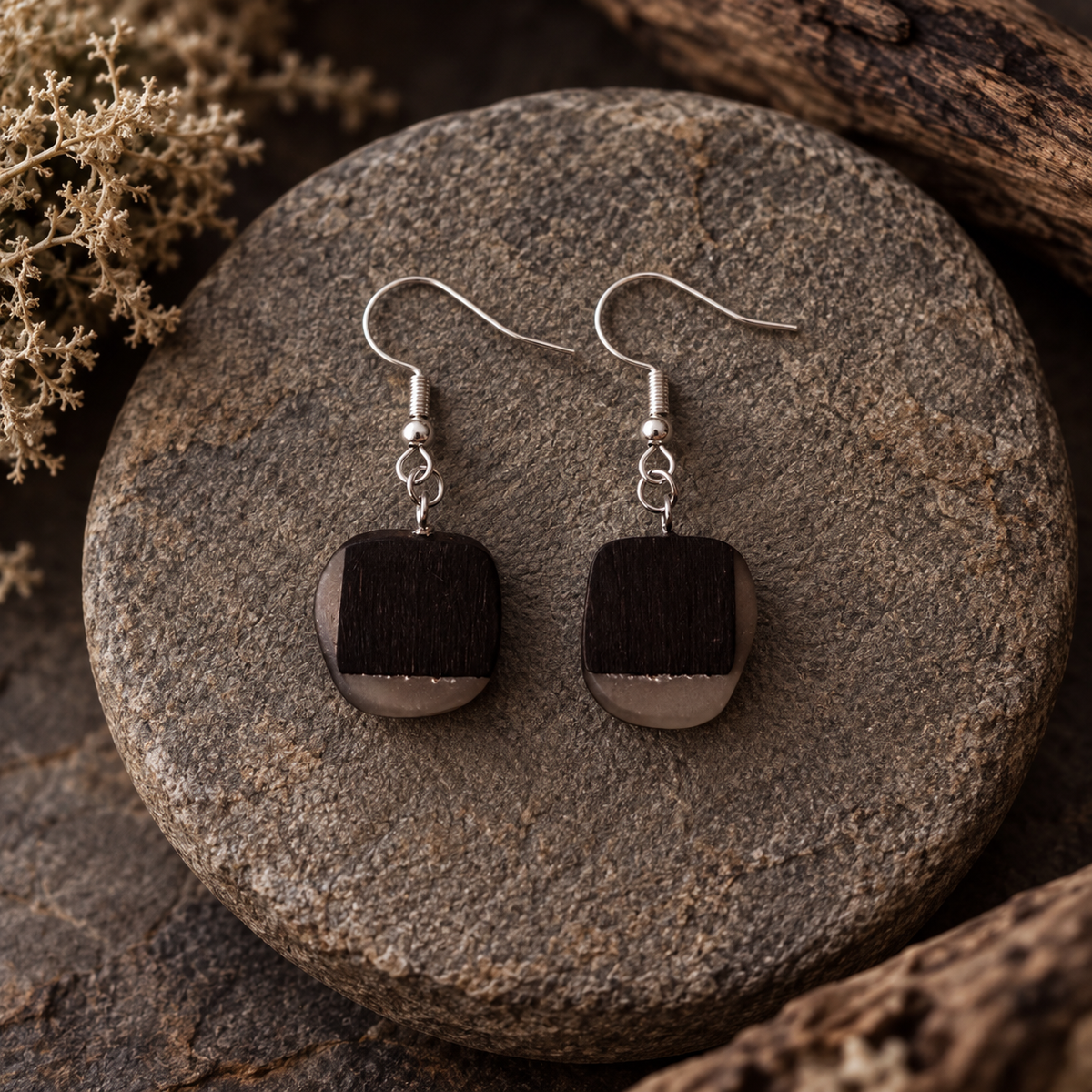 A pair of handmade dangle earrings with square dark brown wooden pendants and silver fishhook wires, displayed on a round gray stone with dried botanicals in the background.