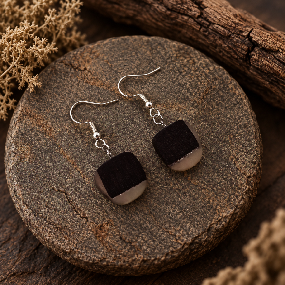 A pair of handmade dangle earrings with square dark brown wooden pendants and silver fishhook wires, displayed on a round wooden slice with dried floral decor.