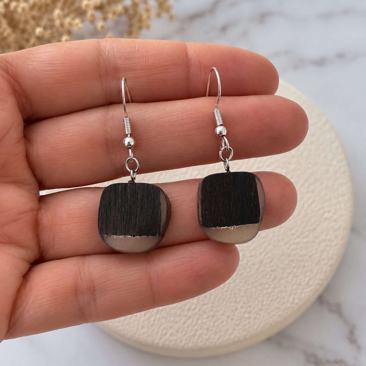 A pair of handmade square drop earrings with a black and silver wood-grain pattern, held in a person's hand over a round textured cream plate.