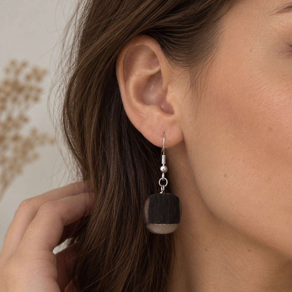 A close-up of a woman's ear wearing a single drop earring with a silver-tone hook and a dark, square-shaped stone pendant that has a silver-toned bottom cap.