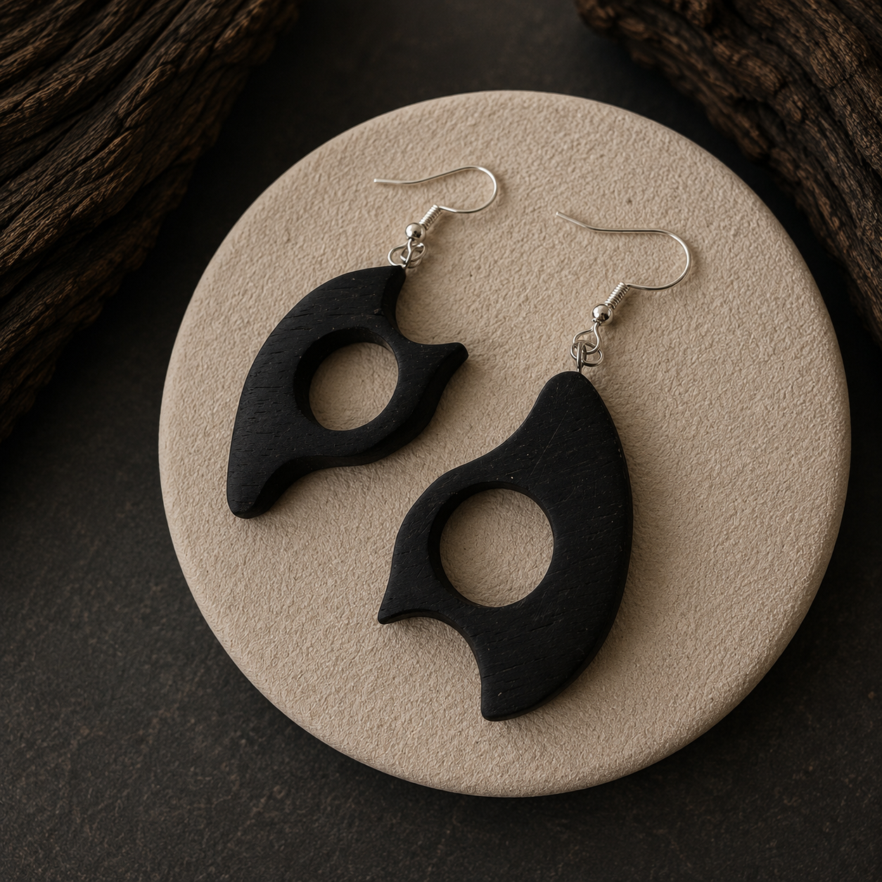 A pair of black wooden dangle earrings with a unique cutout shape and silver fishhook wires, displayed on a round beige stone tray with dark wood in the background.