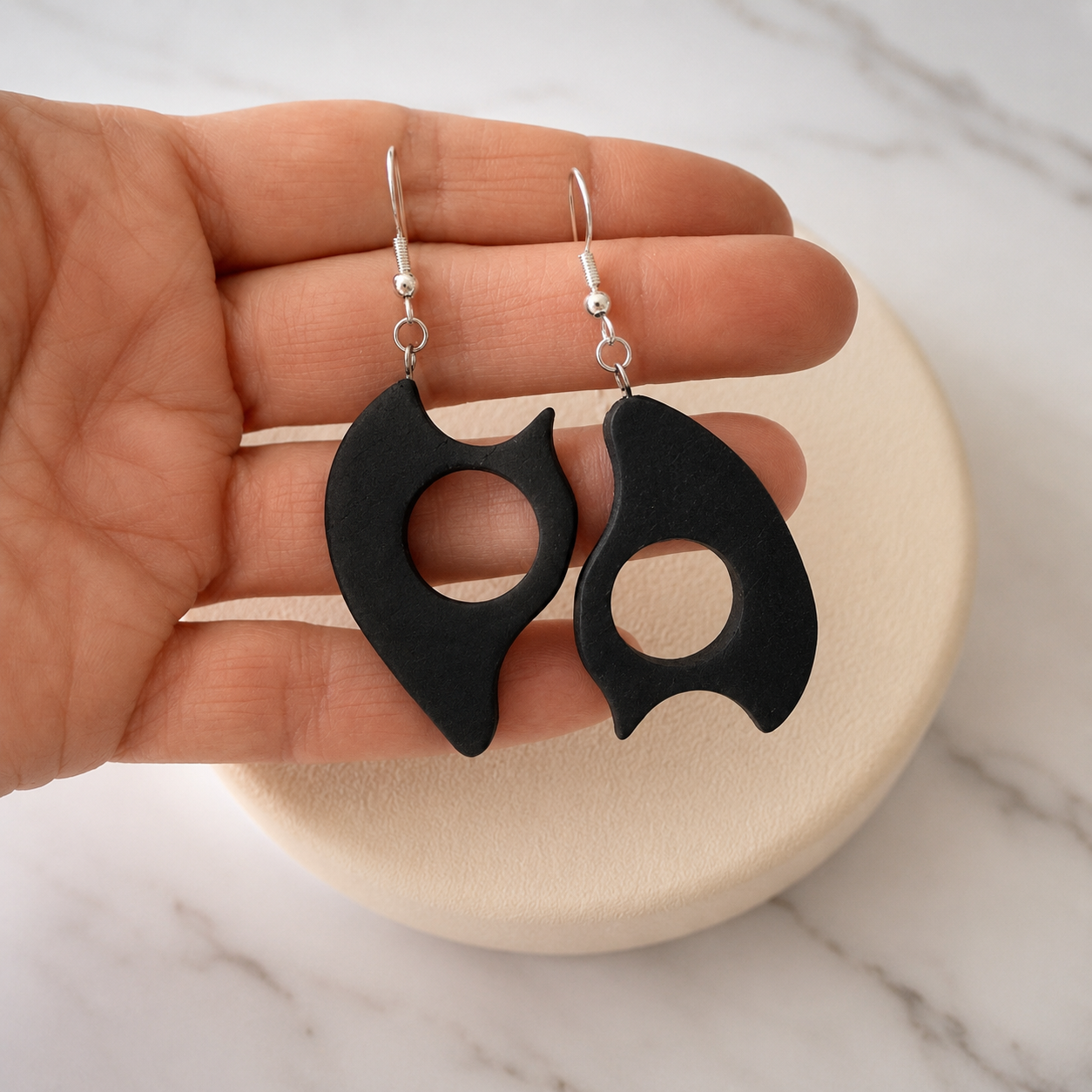 A pair of black geometric drop earrings with silver fishhook wires, held in a person's hand over a round beige display stand on a white marble surface.