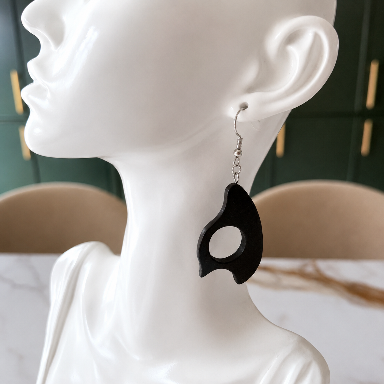 A black organic-shaped dangle earring with a silver fishhook wire is displayed on a white ceramic female mannequin head. The background shows a blurred green cabinet and beige chairs.