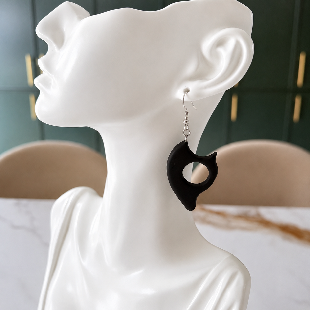 A black organic-shaped dangle earring displayed on a white female mannequin bust, with a dark green paneled wall and beige chairs in the background.
