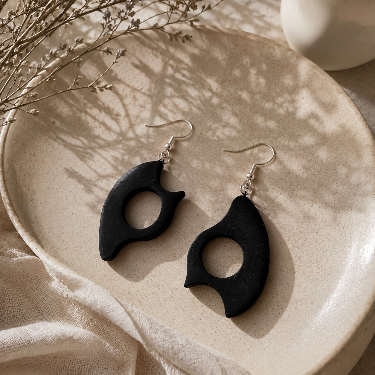 A pair of black organic-shaped dangle earrings with silver fishhook wires, displayed on a beige ceramic plate with soft shadows and dried floral accents.