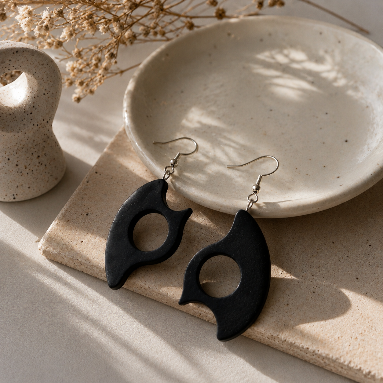 A pair of black wooden dangle earrings with a circular cutout, displayed on a beige stone slab next to a speckled ceramic dish and dried flowers.