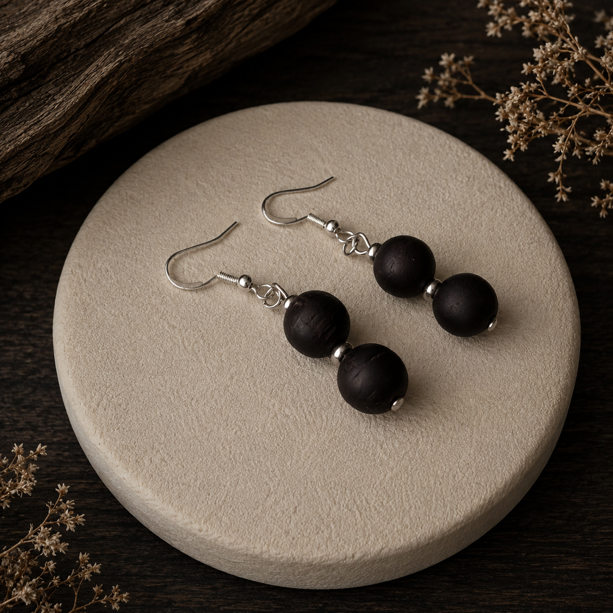 A pair of drop earrings with matte black wooden beads and silver fishhook wires, displayed on a round beige ceramic plate against a dark wood background with dried flowers.