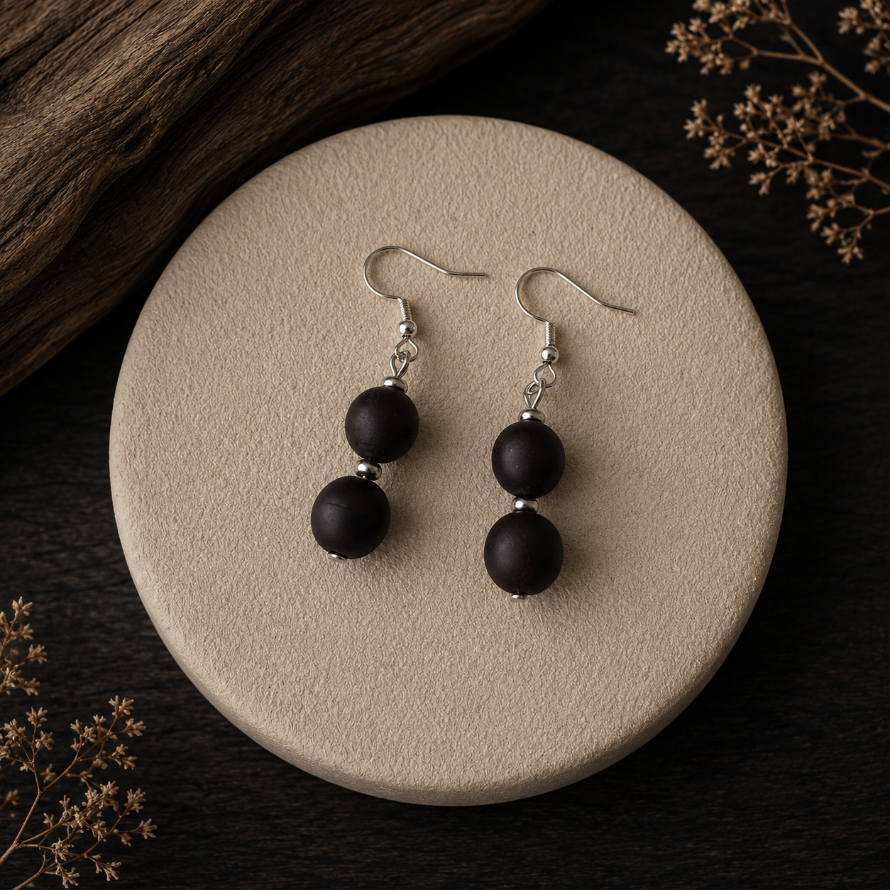 A pair of black beaded drop earrings with silver fishhook wires, displayed on a round beige textured coaster over a dark wood background with dried floral accents.