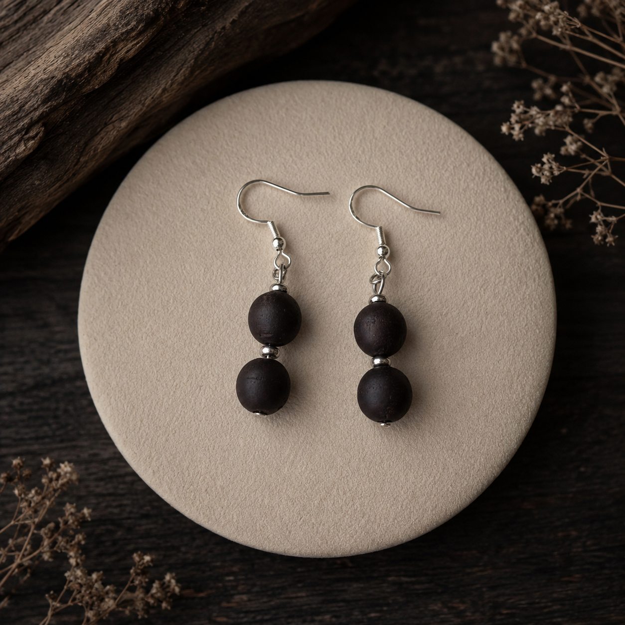 A pair of double-bead drop earrings with matte black beads and silver fishhook wires, displayed on a round beige stone tray with dried flowers and a piece of wood in the background.