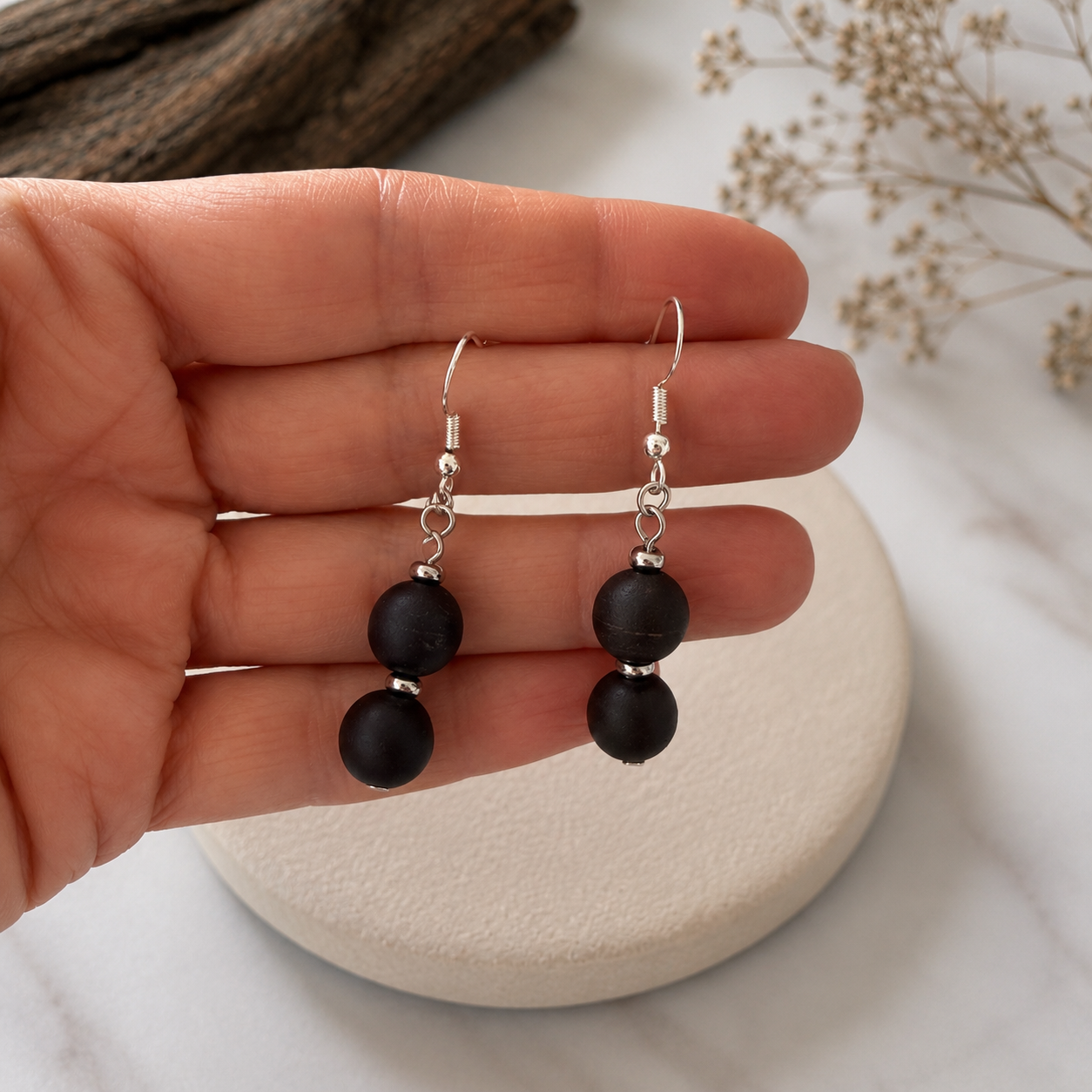 A pair of black beaded drop earrings with silver fishhook wires, held in a person's hand over a round beige coaster on a white marble surface.
