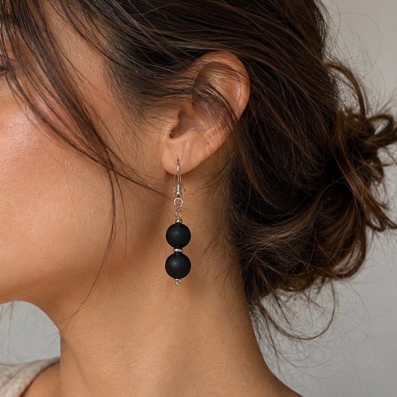 A close-up of a woman's ear wearing a dangling earring with two matte black round beads connected by a small silver spacer and silver fishhook backing.