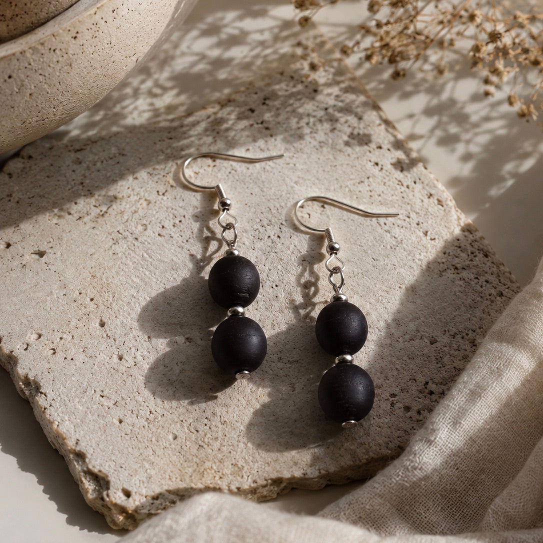 A pair of drop earrings with matte black round beads and silver fishhook wires, displayed on a textured stone slab with linen fabric and dried flowers in soft natural light.