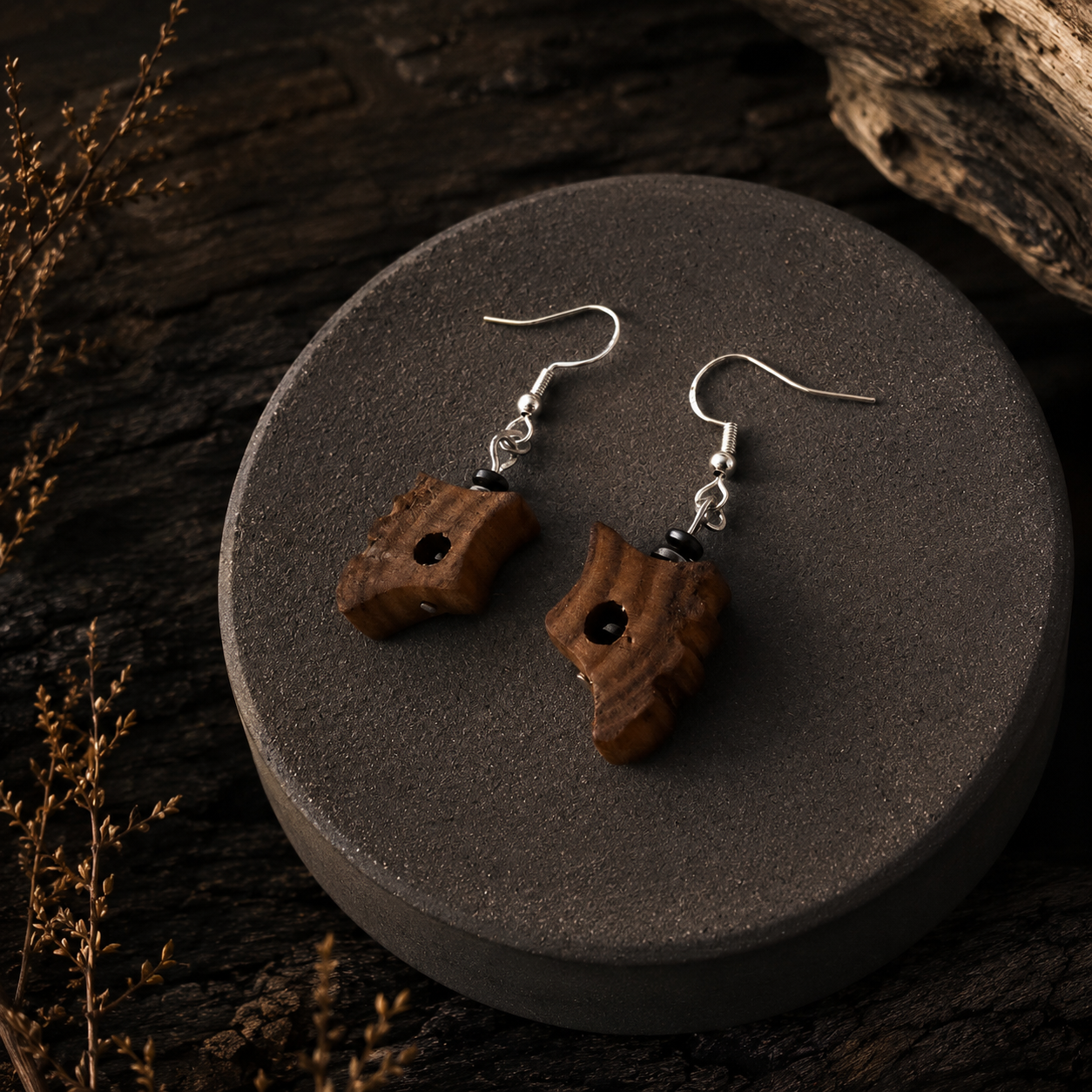 A pair of handmade wooden dangle earrings with silver fishhook wires and small black beads, displayed on a round dark gray stone plate over a rustic dark wood background.