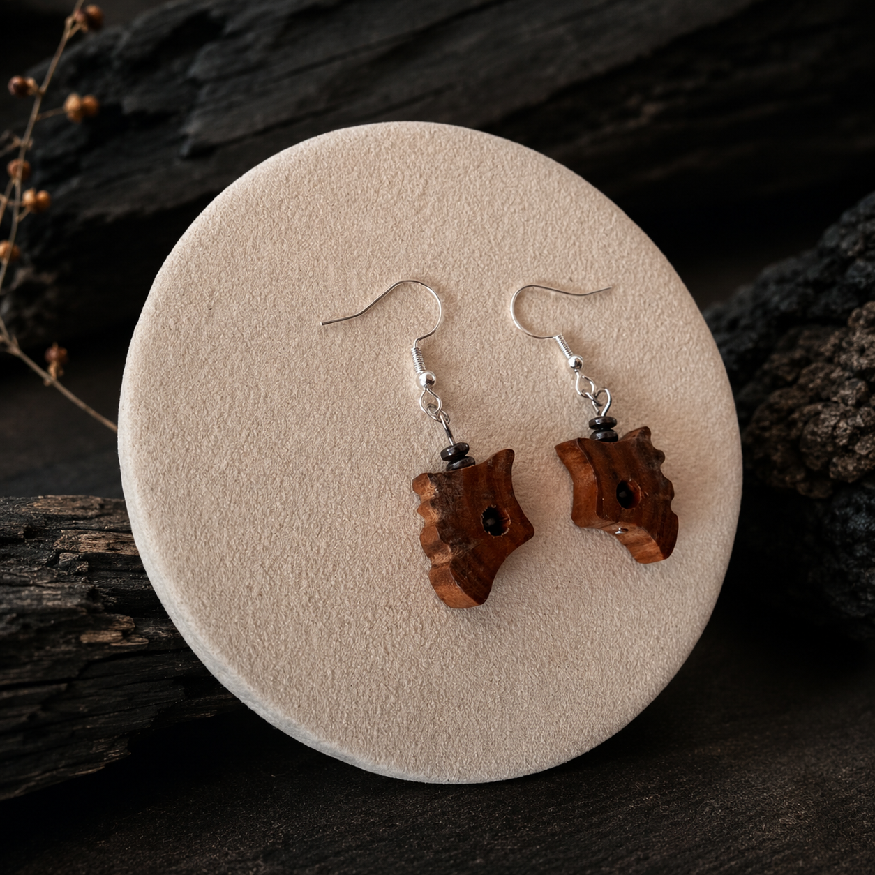 A pair of handmade wooden dangle earrings with silver fishhook wires and small black beads, displayed on a round beige stone stand against a dark rustic wood background.
