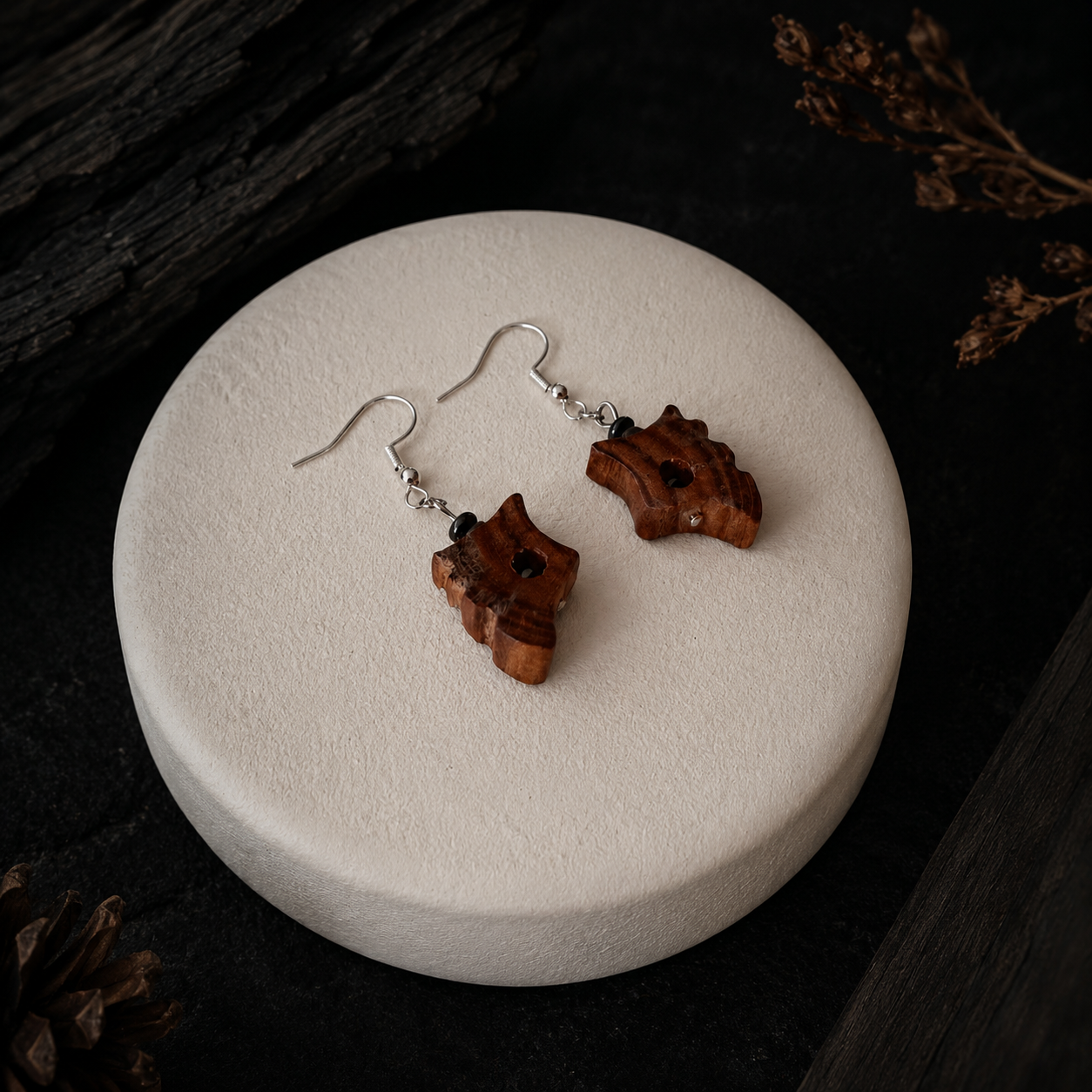 A pair of handmade wooden dangle earrings with silver fishhook wires, displayed on a round textured cream-colored stone tray against a dark rustic wood background.