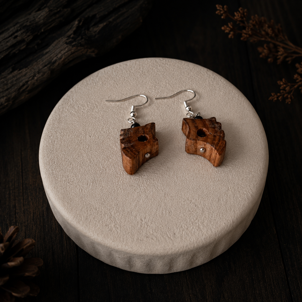 A pair of handmade wooden dangle earrings with carved animal head shapes and silver fishhook wires, displayed on a round textured beige pedestal against a dark wood background.