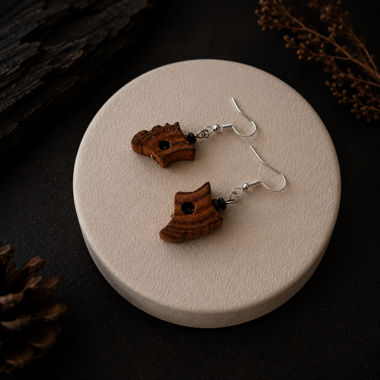 A pair of handmade wooden dangle earrings with carved brown wood pieces and small black beads, hanging from silver fishhook wires, displayed on a round beige pedestal against a dark background.
