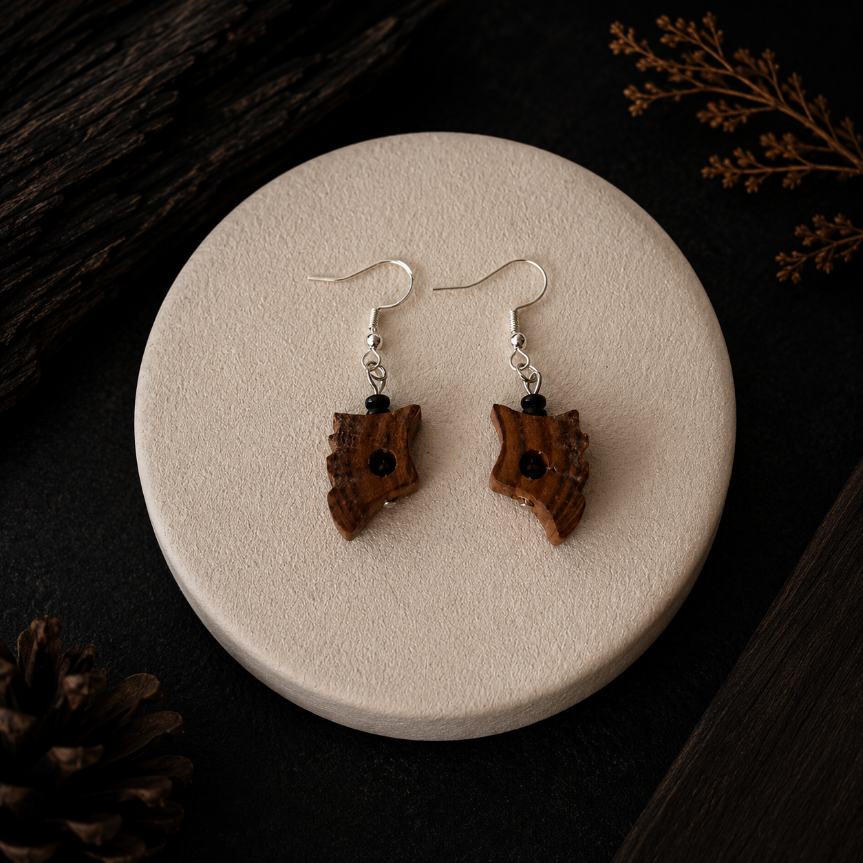 A pair of handmade wooden dangle earrings with silver fishhook wires, displayed on a round beige ceramic plate against a dark wood background with decorative pinecones and dried leaves.