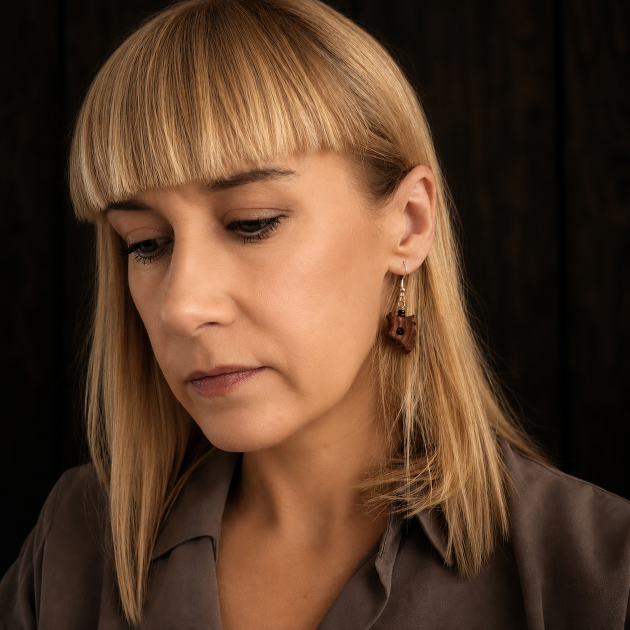 A close-up portrait of a woman with blonde hair and bangs wearing a brown shirt and a dangling geometric earring against a dark background.