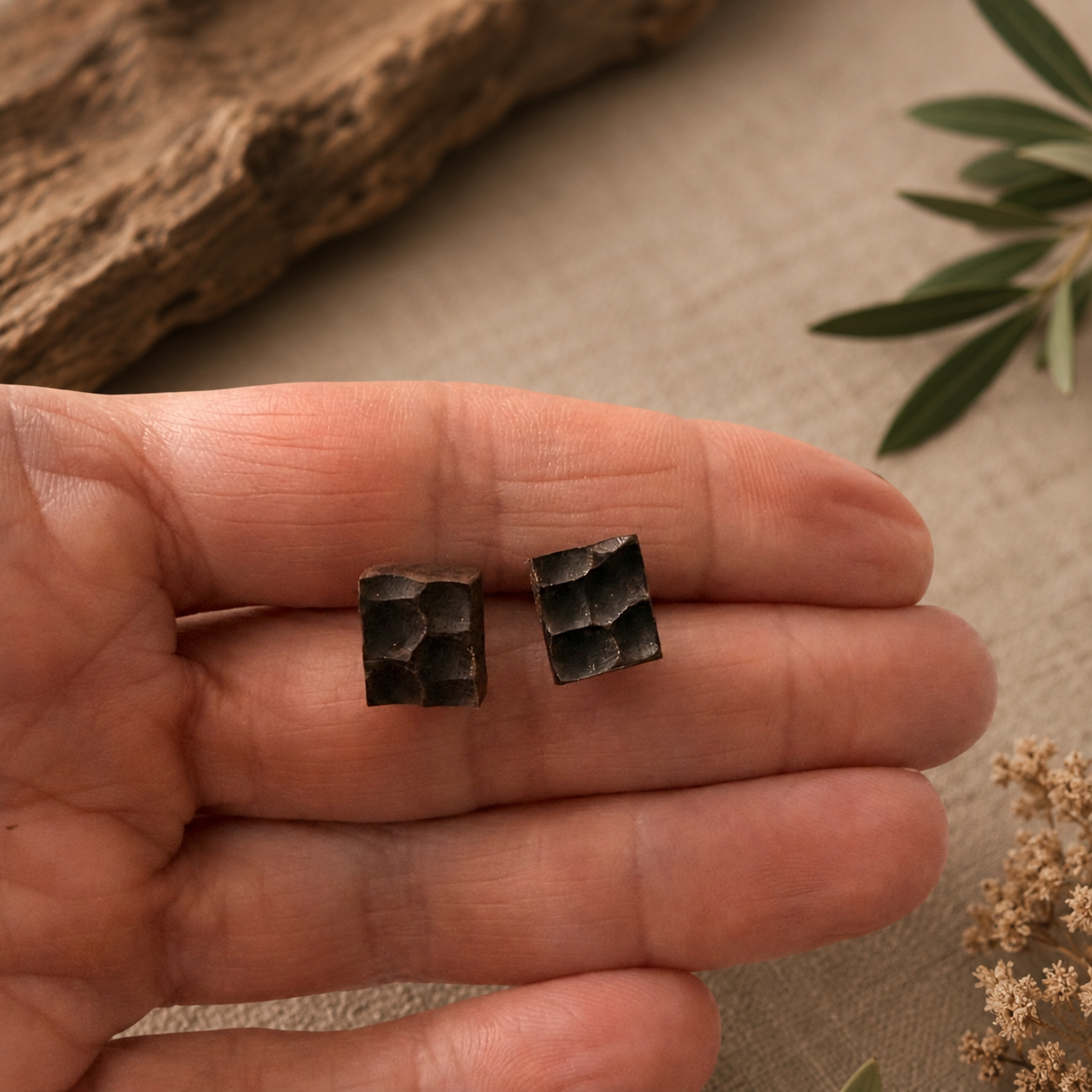 Two small bronze-colored square earrings held between fingers on a neutral background.