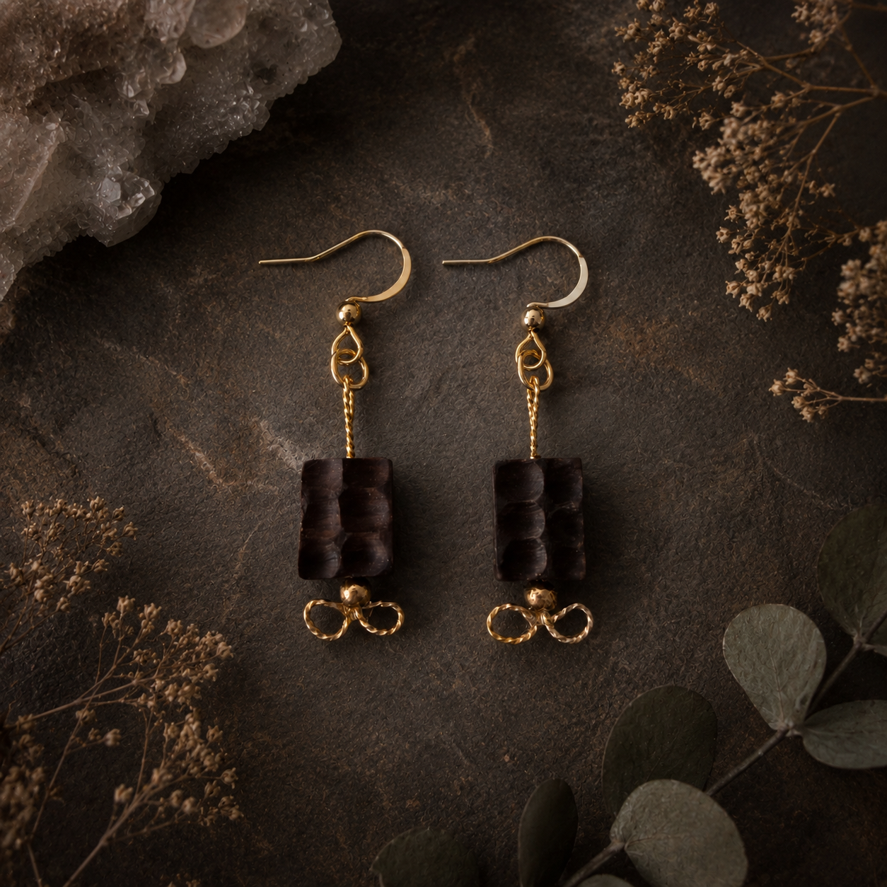 Pair of black earrings with gold accents on a dark textured surface with dried plants.