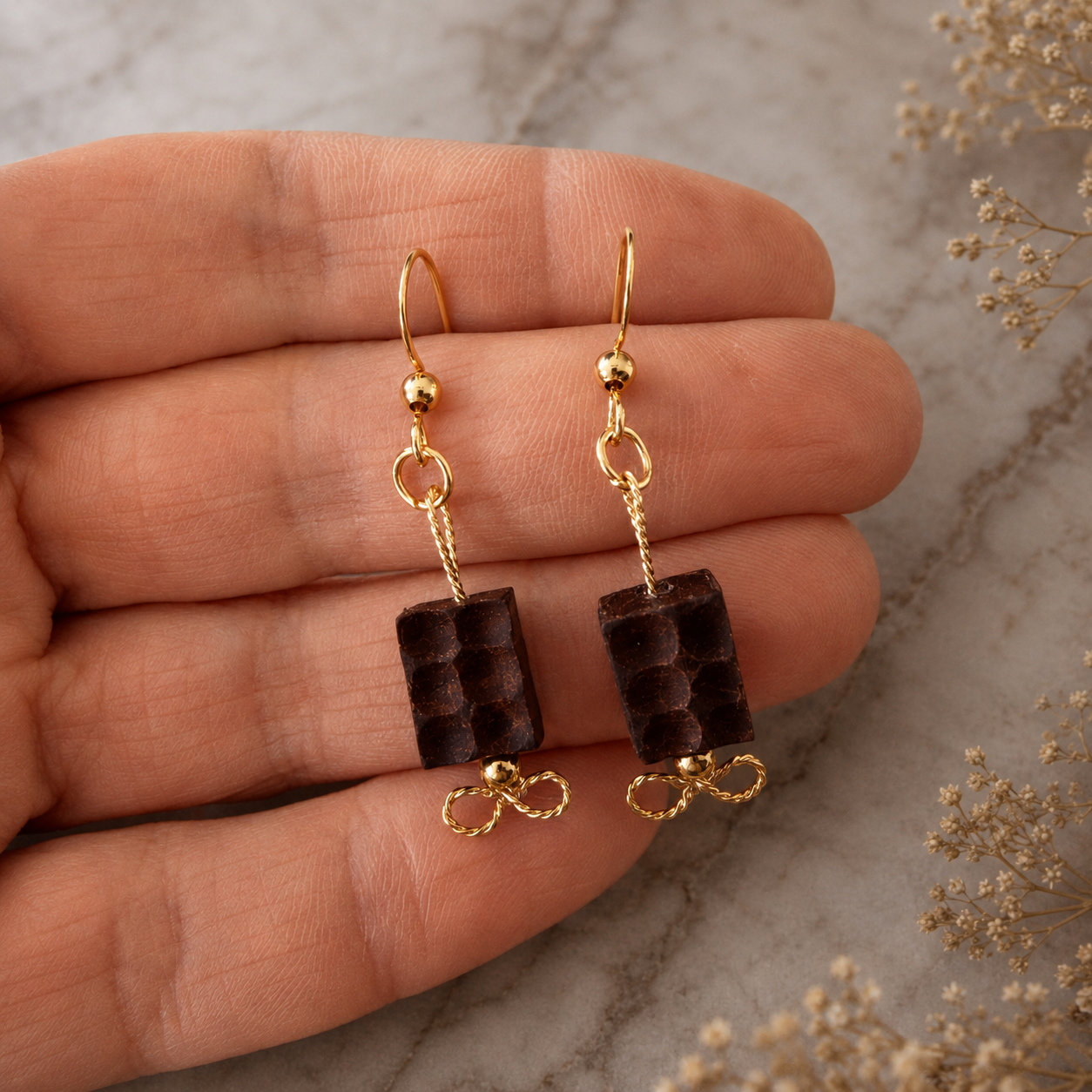 Gold earrings with brown beads held between fingers on a marble surface.