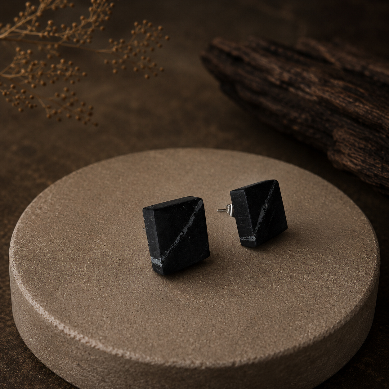 Black square stud earrings on a brown textured surface with dried plants in the background