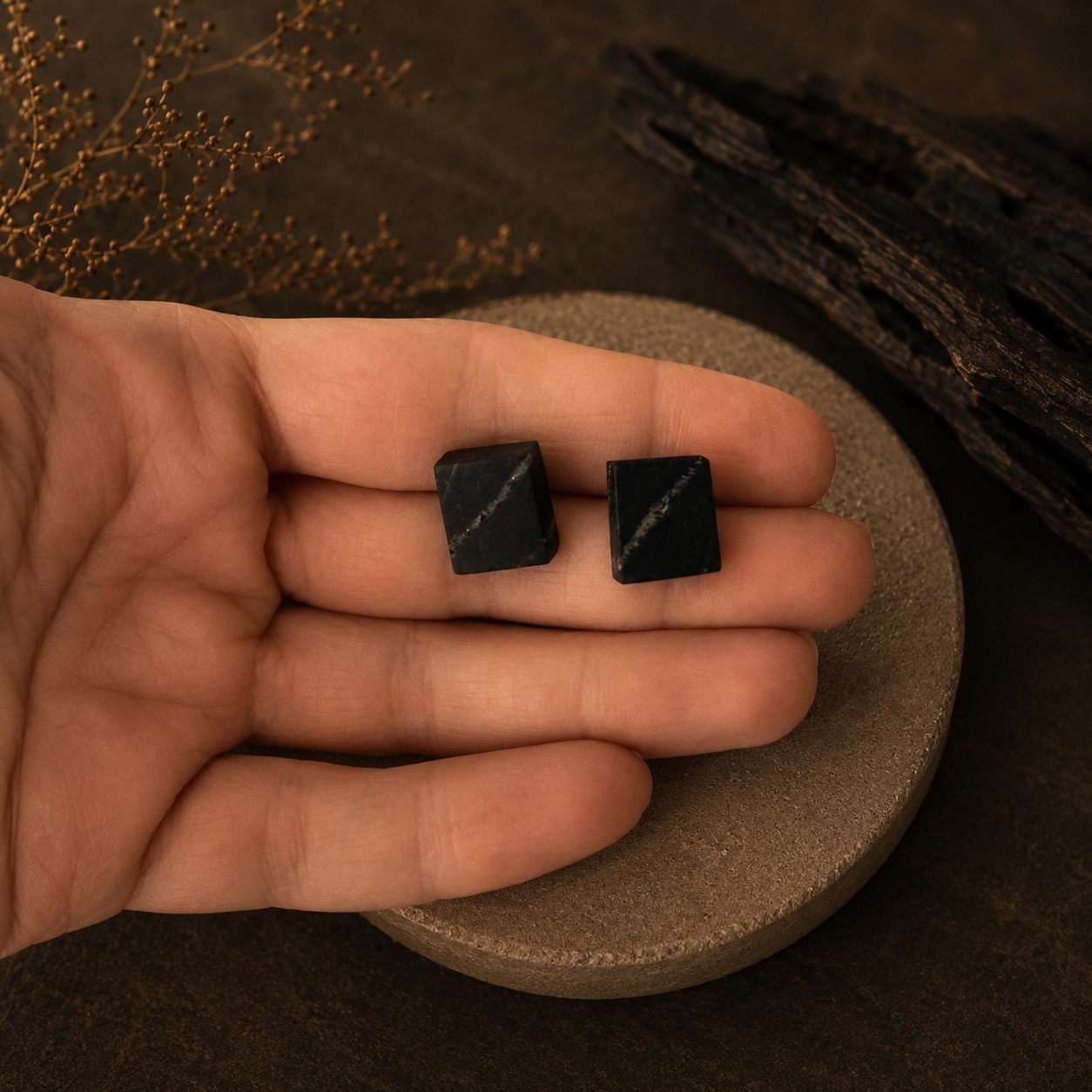 Two black square stones held in a hand on a brown surface with natural elements.