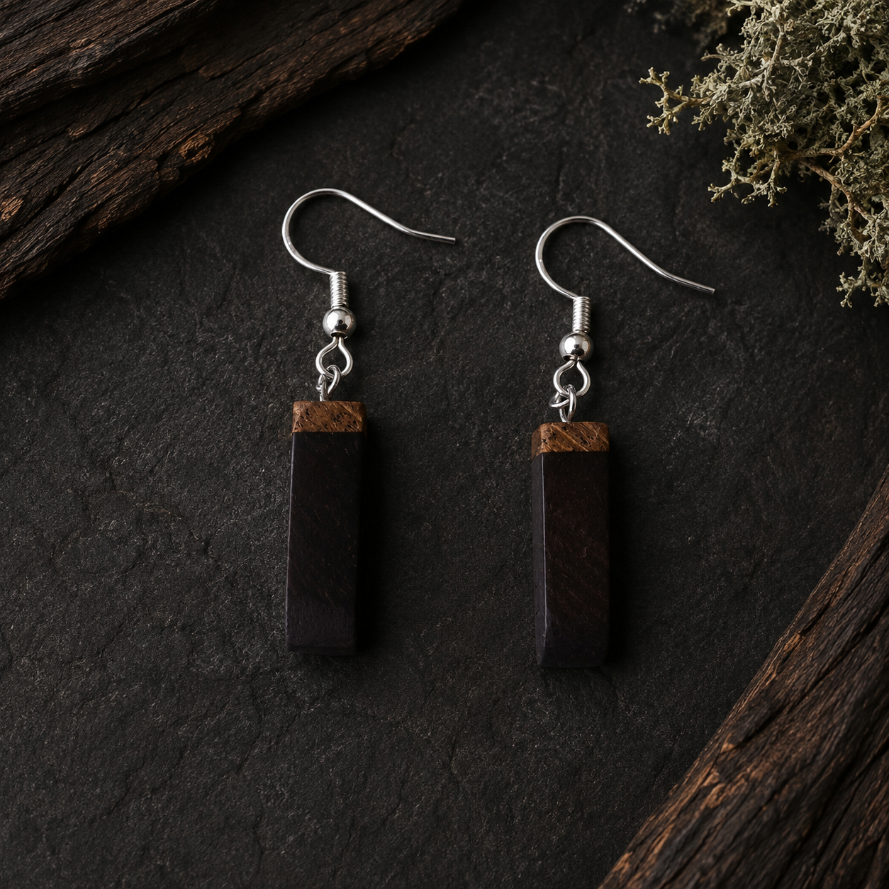 A pair of drop earrings with dark brown rectangular wooden pendants and silver fishhook wires, displayed on a dark textured surface with rustic wood and dried greenery.