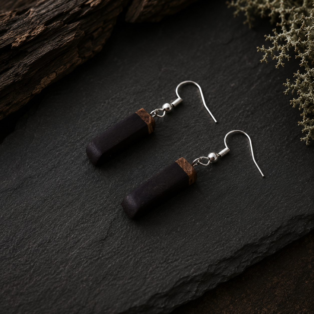 A pair of handmade dangle earrings with dark brown rectangular wooden beads and small tan leather accents, hanging from silver fishhook wires on a dark slate surface.