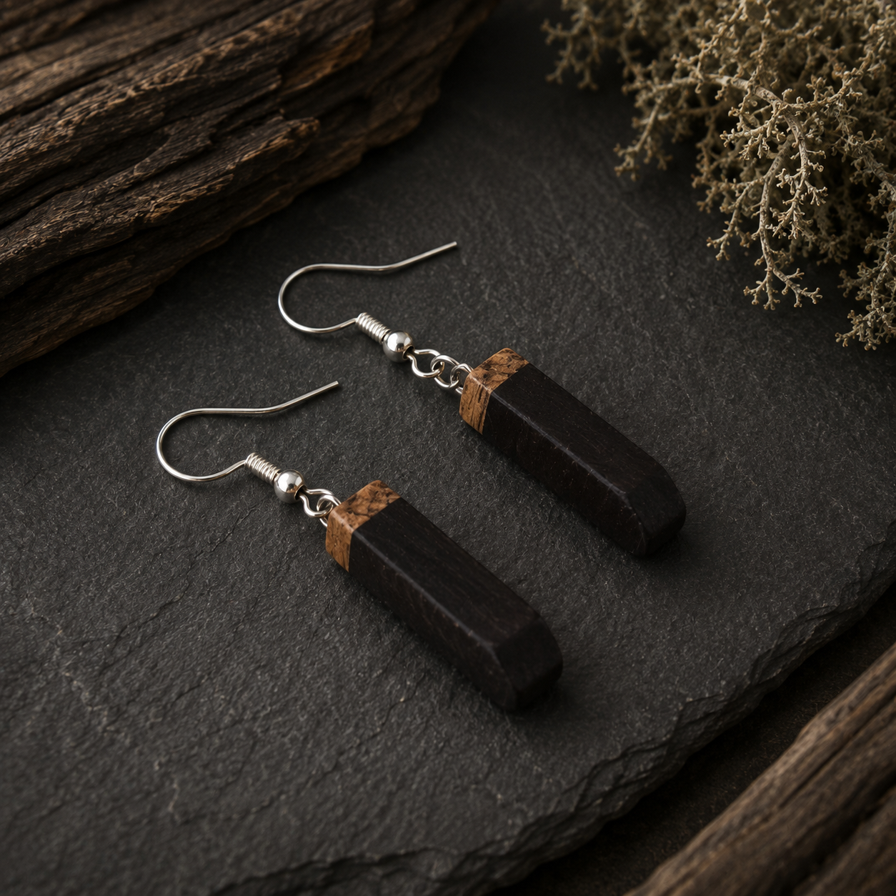 A pair of dark wood drop earrings with silver fishhook wires and a thin brown wood accent band near the top, displayed on a dark slate surface with rustic wood and dried botanicals.