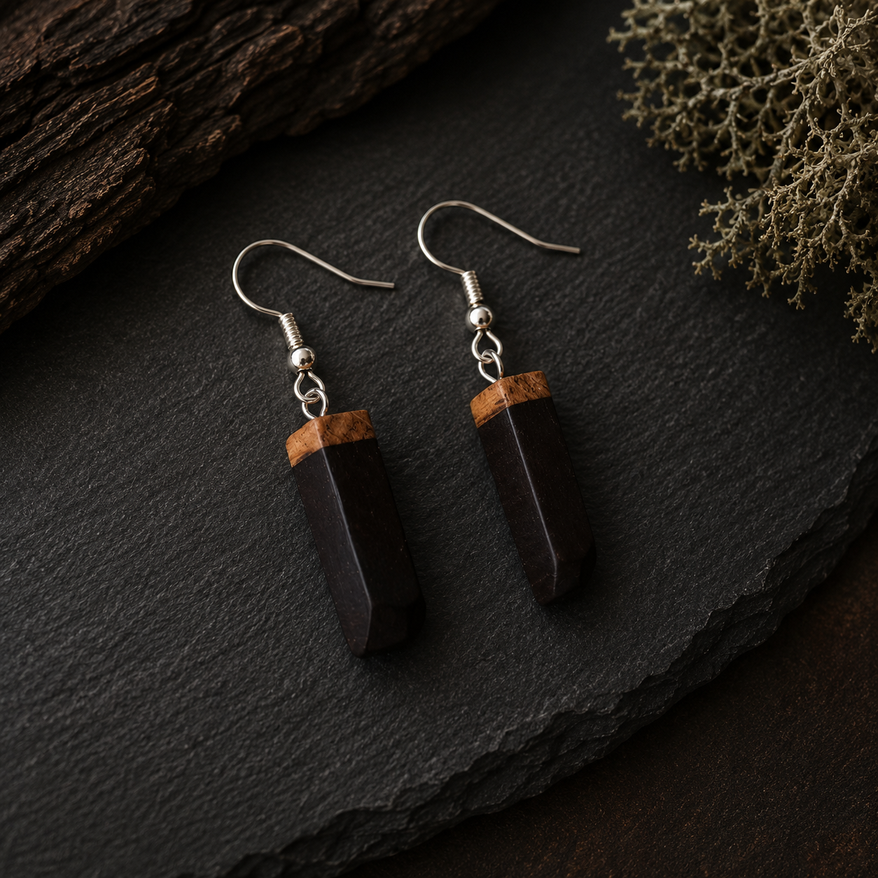 A pair of dark brown wooden pendant earrings with silver fishhook wires, displayed on a dark slate surface with rustic wood and dried botanicals.
